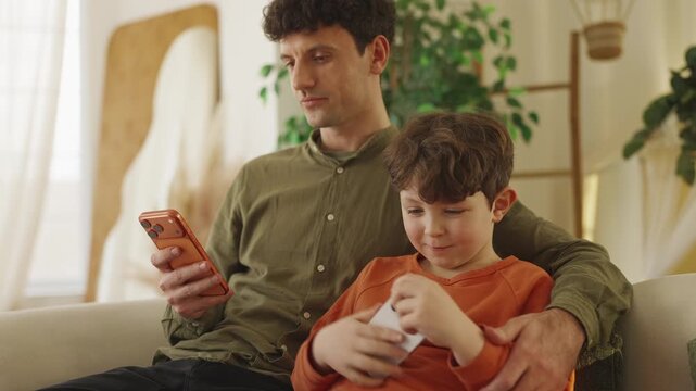 Little boy plays phone video game while his dad messages beside him at home