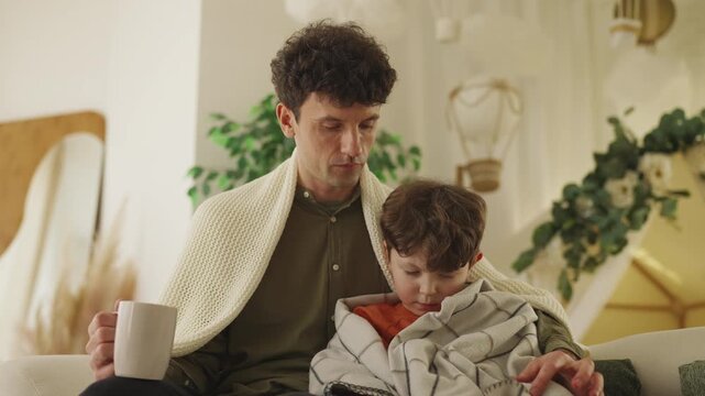 Caring dad hugs sick little son with flu while sitting on sofa at home