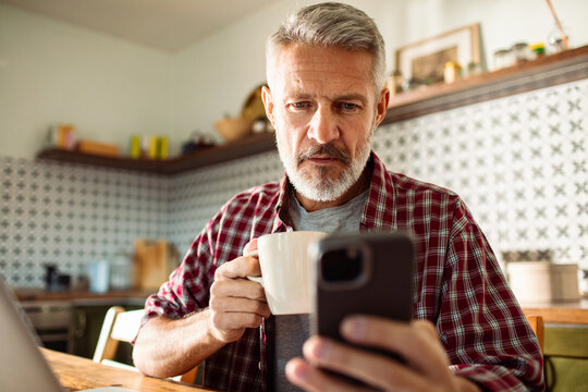 Mature man drinking coffee and checking smartphone in kitchen
