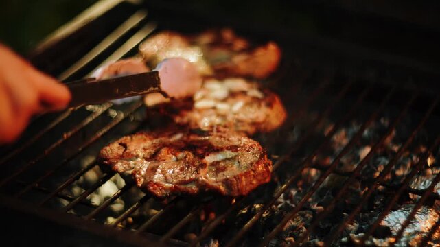 Grilling delicious steaks on hot charcoal at outdoor evening barbecue party with metal tongs