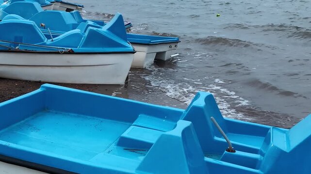Several blue pedal boats rest along the edge of a body of water, with gentle waves lapping against the shore. The boats have molded seats and pedals visible, designed for recreational use.