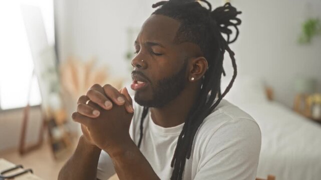 Young african american man with clasped hands praying at a desk in a building; serenity reflection.
