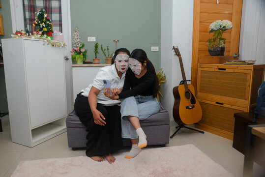 Facial mask woman look at smartphone with friend together for skincare routine, during beauty treatment activity on sofa in cozy home living room for indoor leisure lifestyle concept.
