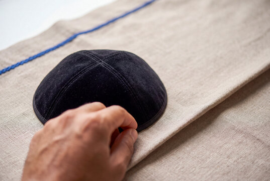 Close-up of a man's hand reaching for a traditional black velvet Jewish kippah or yarmulke resting on a beige linen tallit prayer shawl