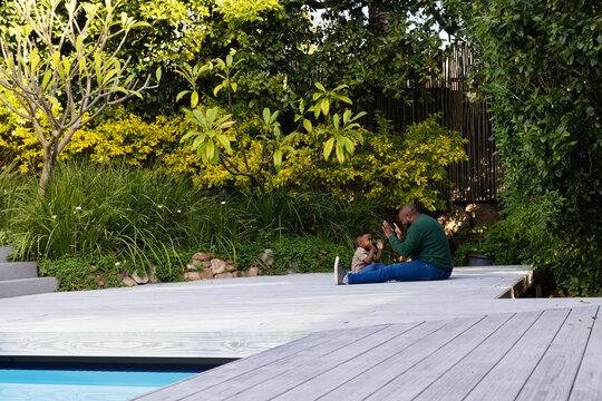 Adult male wearing green top playing clap game alongside youth on deck by pool, copy space