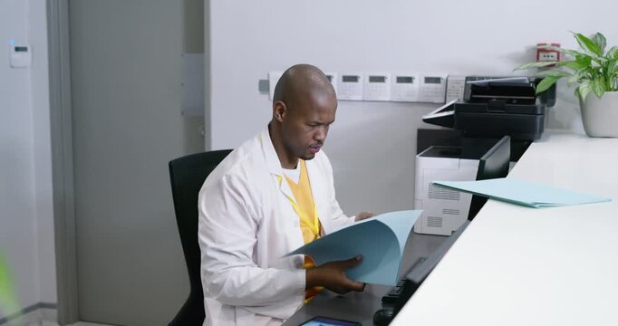 Male medic in lab coat reviewing blue folder, typing patient data and processing forms at reception