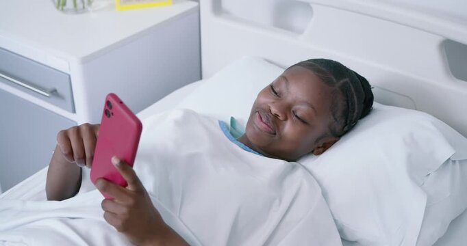 African adolescent female in hospital gown getting message, tapping red smartphone in bed laughing
