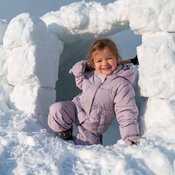 Smiling child in purple snowsuit climbing out of an igloo for winter background and family holiday banner