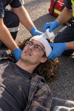 Paramedics bandaging head injury of young man lying on asphalt during emergency roadside treatment