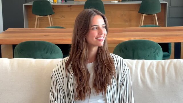 Attractive young woman sitting comfortably on a soft couch inside contemporary coworking space, looking aside with a gentle smile and resting during workday break