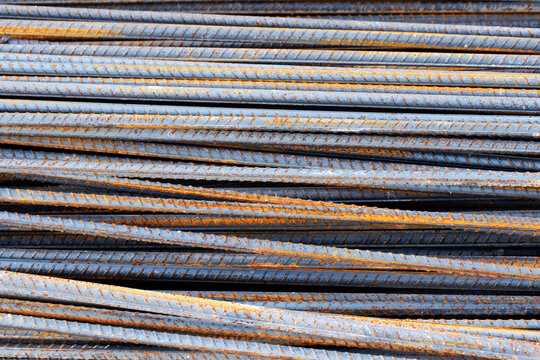 Stacked deformed reinforcing steel rebar rods at a construction site, featuring prominent ribs for concrete bonding and patches of orange surface rust from outdoor exposure.