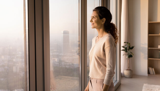 Woman Feeling Hopeful About a New Chapter. South Asian woman standing beside a large apartment window, looking toward the morning skyline with a subtle hopeful smile