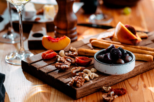 Black olives bowl with walnuts and dried apricots on wooden board - gourmet lunch food pairing concept with white wine glass. Mediterranean appetizers setup for dinner catering.