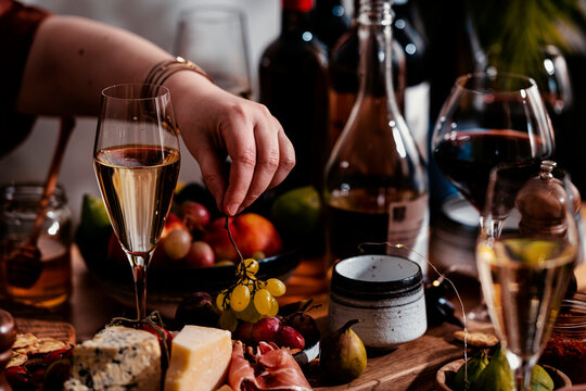 Hand placing green grapes on charcuterie board for wine lunch, gourmet autumn food pairing. Luxury dining experience concept. Flute with sparkling champagne and snacks on table setup.
