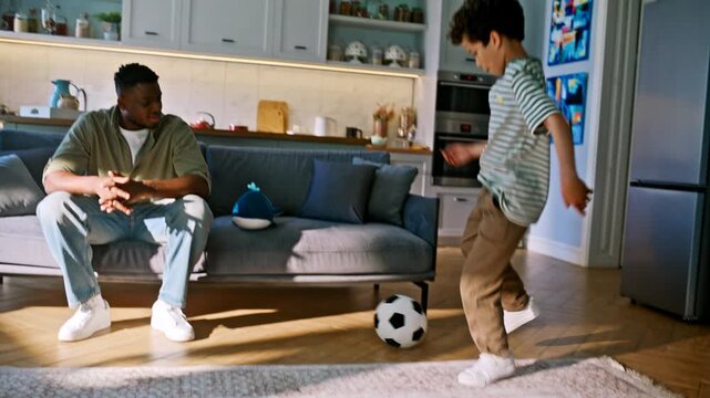 Family playing soccer ball home living room. Boy dribbling, dad cheering couch.