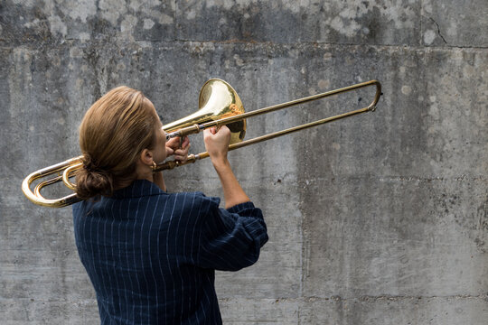 Side profile of a blonde woman playing a gold brass trombone with extended slide outdoors against a stone background