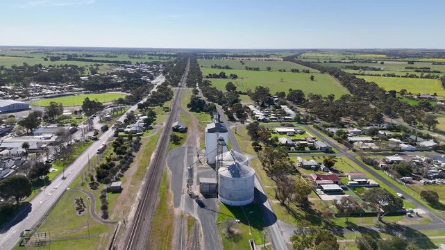 Aerial drone view of Dukes Highway and parallel railway line through Keith in South Australia, with large grain silos and bulk storage facilities beside the train tracks, residential streets.