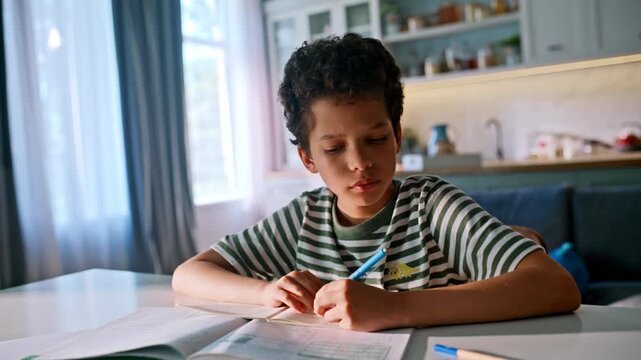 Boy writing homework notebook kitchen table. Focused mixed race pupil smiling.
