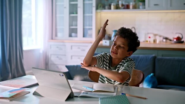 Kid raising hand answering online lesson tablet. Boy doing homework kitchen.