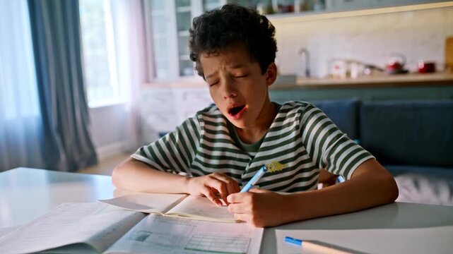 Frustrated schoolboy struggling homework kitchen table. Tired boy holding head.