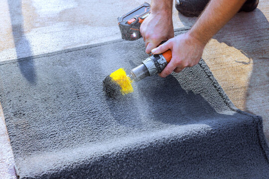Worker uses handheld powered cleaner car carpet to treat carpet on paved surface