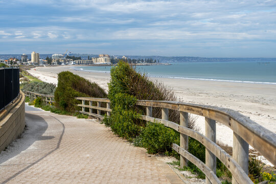 A paved coastal shared path curves along the foreshore at West Beach in Adelaide, South Australia, Gulf St Vincent shoreline stretching toward the apartment towers and skyline of Glenelg North.