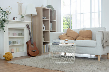 Bright cozy living room interior with a gray sofa, beige pillows, acoustic guitar, bookshelf, and round coffee table by a large sunlit window, featuring modern home decor and relaxing atmosphere. © Ratirat
