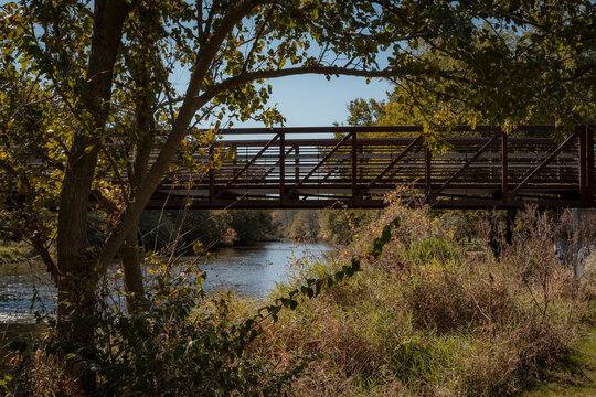 Metal footbridge spanning a scenic river surrounded by autumn trees and lush foliage on a sunny day