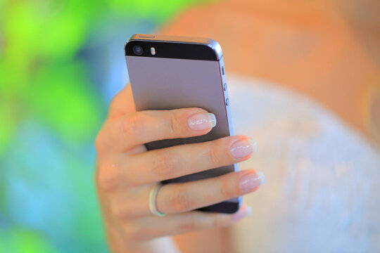 Close up of a female hand with french manicure holding a grey metallic smartphone on a blurred green background