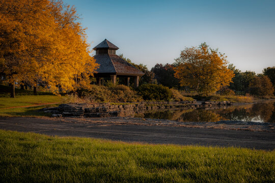 Wooden gazebo shelter surrounded by golden autumn trees along a scenic pond in a quiet public park