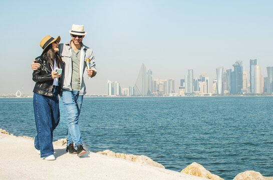 Doha Qatar tourist couple walking Corniche waterfront West Bay with skyline skyscrapers background happy tourists vacation lifestyle Middle East urban city exploration sunny summer fun day bonding