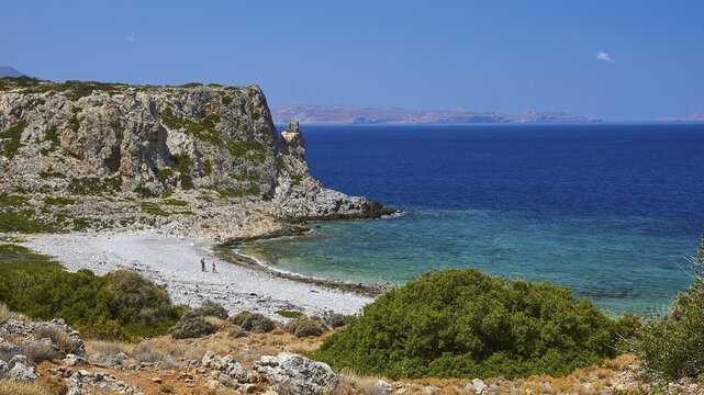 Agios Pavlos beach, pebble beach, barren rocks, trees, blue sea, green sea, Gramvoussa peninsula, two walkers on the beach, almost cloudless blue sky, single white cloud, west coast of Rodopou peninsula, west Crete, island of Crete, Greece