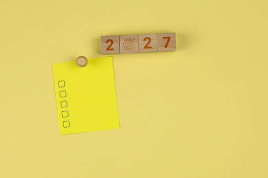 The year 2027 displayed on wooden blocks, accompanied by a target icon. A yellow sticky note features a blank checklist. This represents a concept of strategy, focus, and goals to be achieved.
