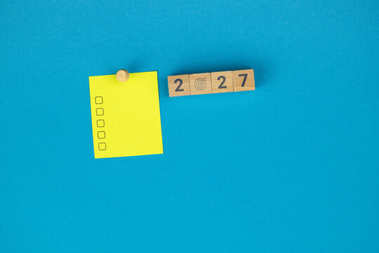 The number 2027 formed with wooden blocks, along with a target icon. A yellow sticky note with empty checkboxes. A concept for new year goal planning. Light blue background.
