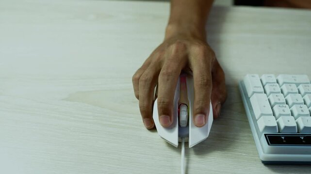 Hand using a computer mouse beside keyboard, Close-up of a hand using a white computer mouse beside a compact keyboard on a light wooden desk.