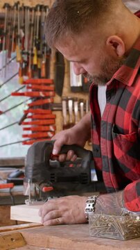 Vertical video. Woodworking process. Using jigsaw. Bearded male carpenter in plaid shirt cutting plank in smoky workshop with professional electric equipment.