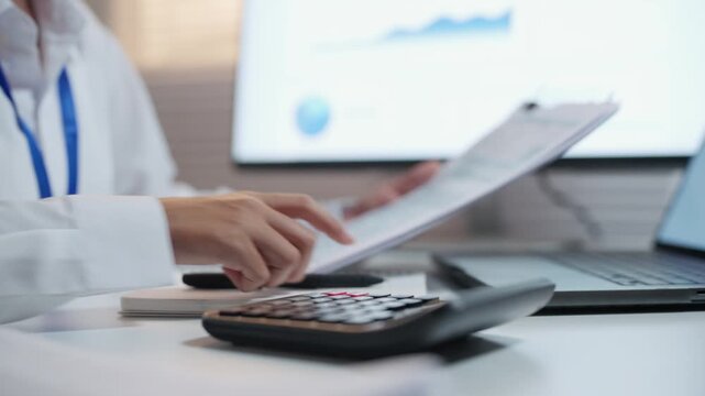 Office worker using a calculator while reviewing printed financial documents at a desk with laptop and charts, illustrating expense calculation and budget review.