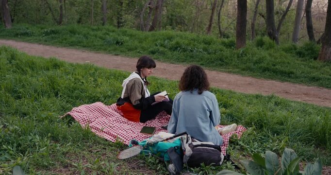 Two young women on red gingham blanket in park clearing with backpacks books and badminton racket while one reads notebook and other listens sitting on grass by dirt path in spring woods scene