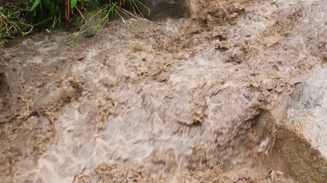 Floodwater Rushes Through Muddy Channel, Turbulent Rapid Stream Erosion Over Rocky River Ground During Severe Rainfall, Powerful Brown Water Torrent, Nature Flood Hazard Scene with Wet Soil and Dense