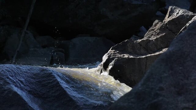 Rocky Stream Waterfall Flowing Through Mountain Gorge, Sunlight Illuminating Textured Water, Rugged Boulder Formations, Deep Shadow, Dramatic Nature Landscape Contrast and Natural Motion
