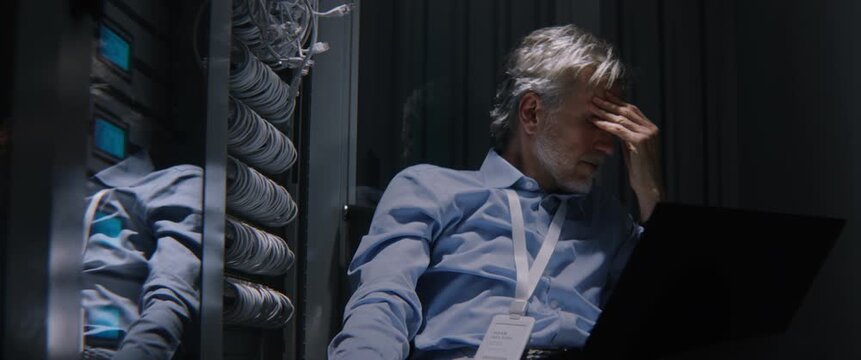Tired Man Sitting Near Server Cabinets, Inspecting Metrics on Laptop, Running Diagnostic Software. Fatigue Meets Critical Infrastructure Work, Enterprise Network Auditing, System Modernization.
