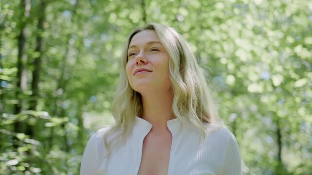 Woman with blonde hair enjoys sunlight in forest. She smiles, eyes closed, soaking in peace. White shirt contrasts with green trees. Ideal for wellness, nature, or relaxation themes