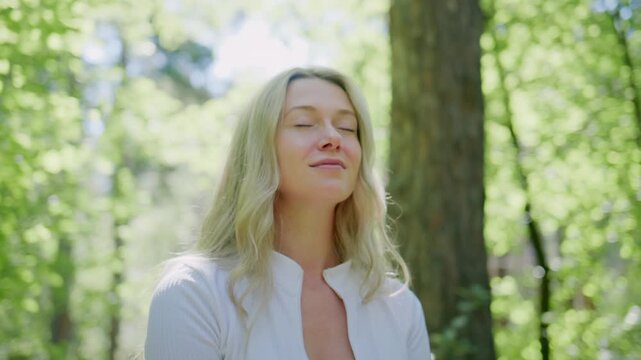 Woman stretches arms upward in sunlit forest. Her eyes close, face relaxed, enjoying calm. Soft light filters through green leaves. She wears white top, standing between tall trees