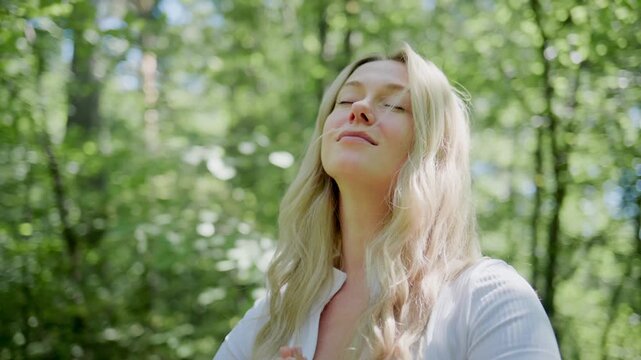 Young woman meditates peacefully in sunlit forest. She smiles softly with eyes closed. Hands folded gently at chest. Calm, serene, and joyful emotion shown. Ideal for wellness, mindfulness