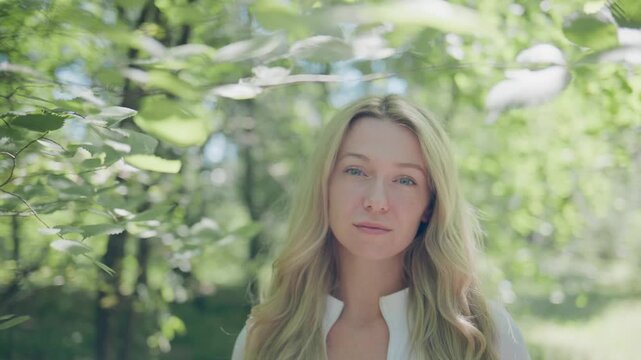 Woman with long blonde hair stands in sunlit forest. Leaves partially obscure her face, creating soft, dreamy framing. She looks calm, thoughtful, slightly serene