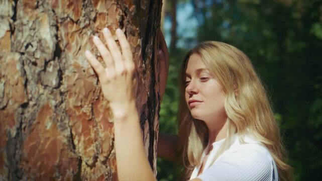 Serene woman gently touches a large pine tree trunk. She leans close, eyes closed, absorbing forest calm. Sunlight filters softly through green foliage. Peaceful moment evokes nature connection