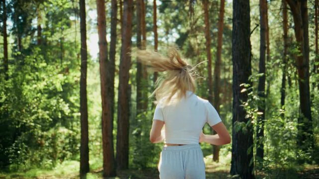 Blonde woman runs through sunlit pine forest. Her hair flows freely as she moves. Scene captures active, peaceful outdoor exercise. Ideal for health, wellness, or nature-themed projects
