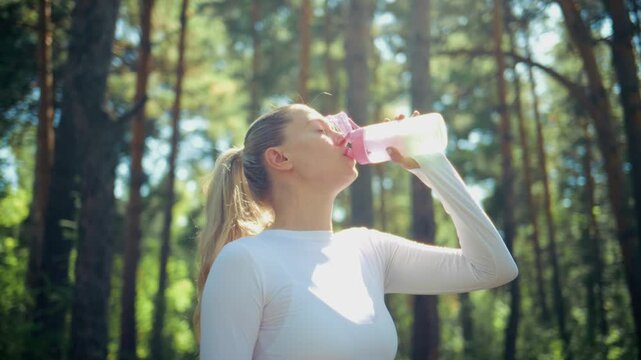 Young woman drinks water in forest after workout. She looks relaxed and refreshed. Sunlight filters through tall pine trees around her. Scene shows post-exercise hydration and nature connection