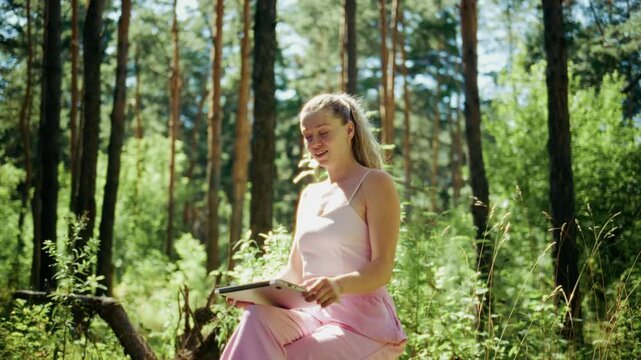 Young woman sits in forest, smiling warmly. She holds book, waves gently toward camera. Sunlight filters through tall pine trees, creating soft light. Scene feels calm and peaceful