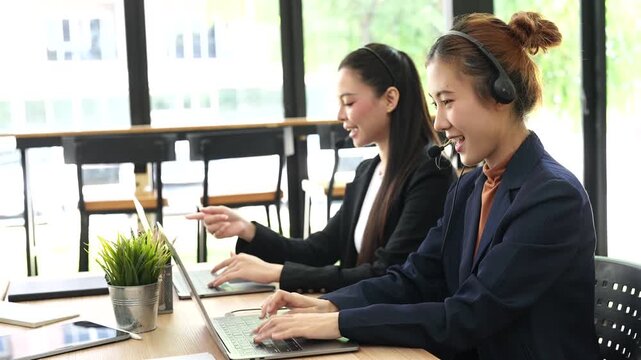 Two young Asian female call center agents wearing headsets, typing on laptops and talking with customers at office desk.